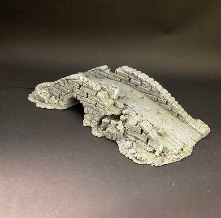 A miniature grey stone bridge or ruined archway terrain piece, likely for tabletop wargaming or diorama use, photographed on a dark grey background. Large pre-painted damaged bridge tabletop gaming terrain scenery