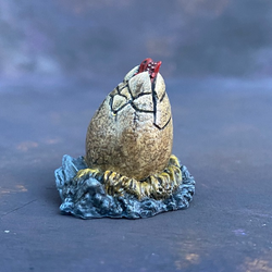 A Mantic Games objective marker in the style of a baby red dragon hatching from its egg. This cute little miniature has a red dragon snout, cracked egg on a bed of hay and stones.