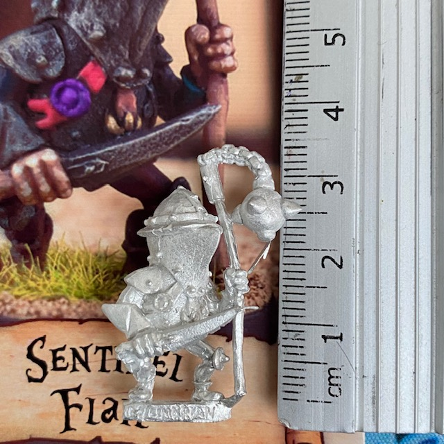 Sentinel Flail by Northumbrian Tin Solider from the Nightfolk sentinel range wears a battered helmet and various other pieces of armour while carrying a flail in one hand and a blade in the other, proudly displaying a ribbon