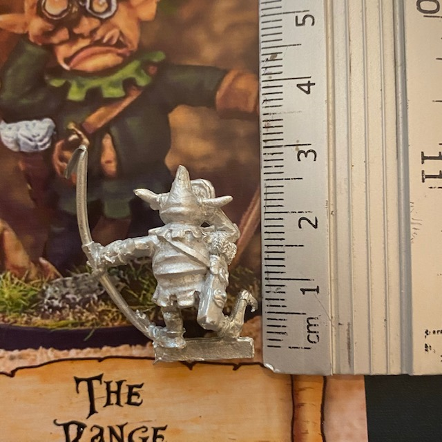 The Range by Northumbrian Tin Solider holds a bow in one hand and a spyglass to his eye with the other hand, with large ears and large floppy front shoes, shown with a ruler