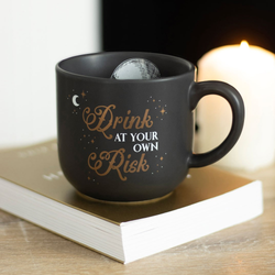 A black mug with a white skull pattern inside and the words Drink At Your Own Risk in gold &amp; white written on the outside with moon and stars showing everyone not to use your mug.