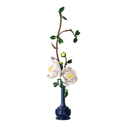 A decorative floral arrangement featuring white magnolia-style flowers with yellow centres, green leaves, and a brown branch, set in a tall dark blue vase against a plain white background. CaDA ARTIZAN EASTERN FLORAL ART building brick floral display