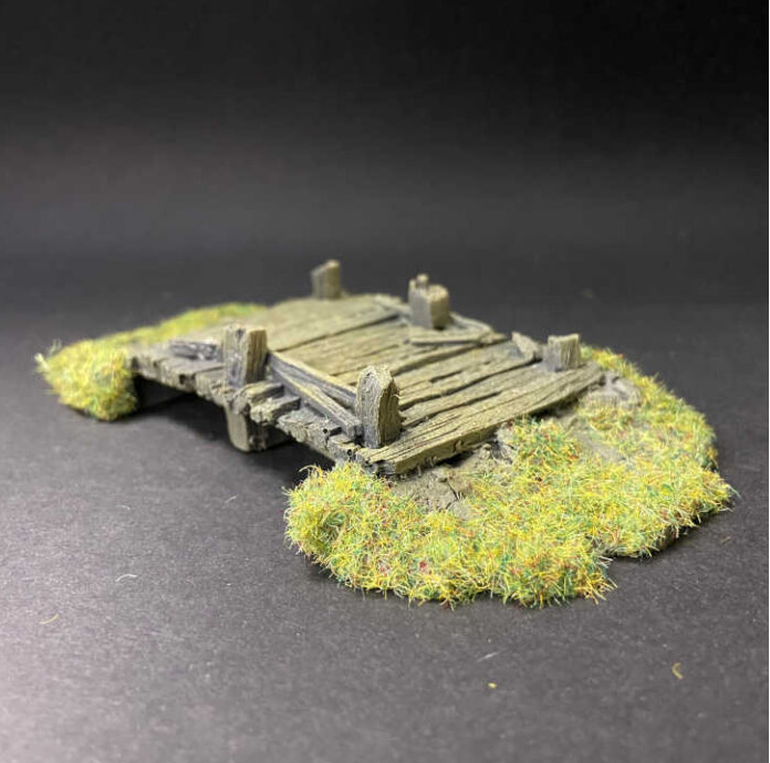 A miniature model of a weathered wooden bridge with green-tinted grass surrounding it, displayed on a dark grey background. Large pre-painted timber bridge tabletop gaming terrain scenery