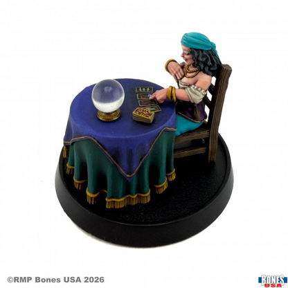 A painted tabletop miniature figurine of a woman seated at a draped table with a crystal ball and tarot cards, mounted on a round black display base against a white background. Madam Estrella Fortune Teller