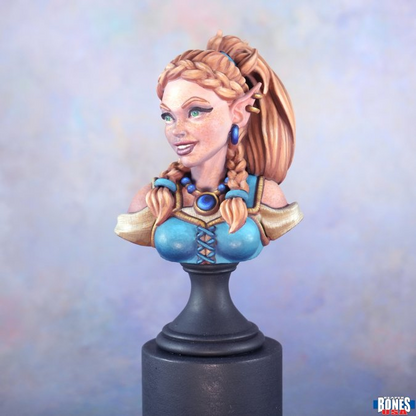A painted fantasy elf bust sculpture on a black display base, featuring long braided light brown hair, blue eyes, a blue and gold outfit, and blue earrings, photographed against a soft pastel blue background. Ryelle Rainheather Bust