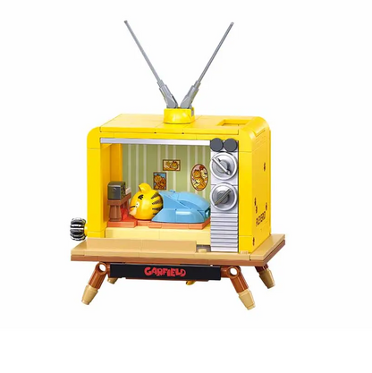 A toy building block set of a retro television set, featuring a bright yellow body, silver dials, wooden legs, and a small interior scene with a yellow bear and blue blanket, shown on a plain white background. Sluban Garfield TV 193 piece brick building kit with sleeping Garfield diorama