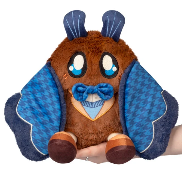 A brown plush toy shaped like a cartoon bat or creature with large blue and white eyes, blue wing-like side panels, a blue bow tie, and blue antler-like tops, being held by a person's hand against a white background. Squishable Academia Mothman