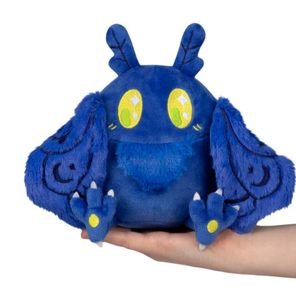 A blue plush toy with large yellow eyes and wing-like ears, being held in a person's hand against a plain white background. Squishable Blue Mothman - Snackers S1E3