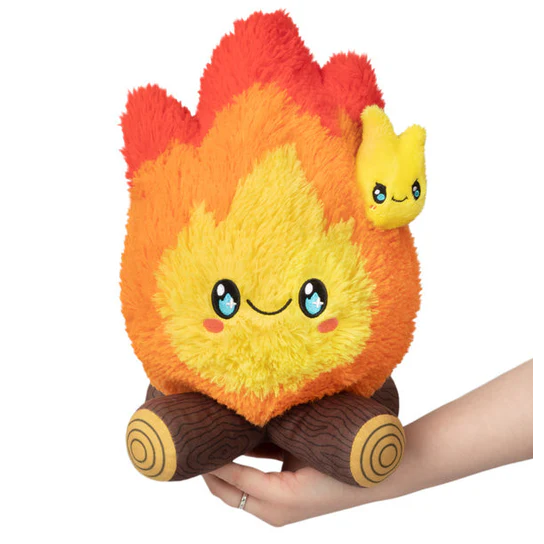 Plush toy shaped like a flame with a small yellow creature on top, held by a hand against a white background. Squishable Campfire