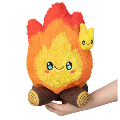 Plush toy shaped like a flame with a small yellow creature on top, held by a hand against a white background. Squishable Campfire