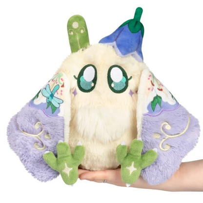 A plush toy shaped like a small fantasy creature or bird, held in a person's hand against a plain white background. The plush has cream-colored fluffy body feathers, large green and blue eyes, lavender wings with floral embroidery, and green feet. Squishable Fairy Mothman