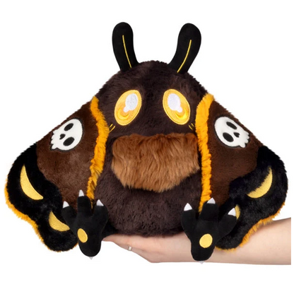 A plush toy shaped like a cute dark brown creature with large yellow eyes, black ears and claws, and white skull details on the wings, being held in a person's hand against a white background. Hawkmothman
