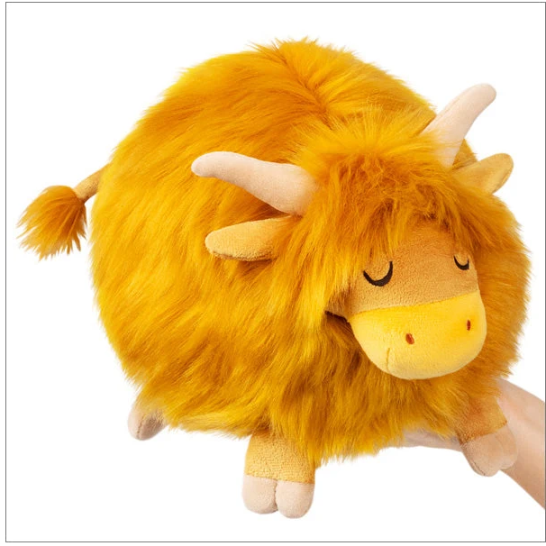 A fluffy yellow-orange plush animal toy with cream horns and a cream snout, shown against a plain white background. A human hand is visible underneath the toy, supporting it. Squishable Highland Cow