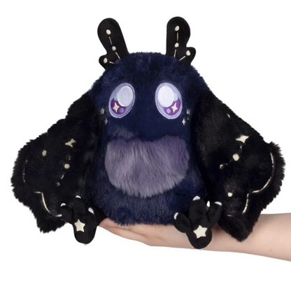 A plush toy shaped like a cute fantasy creature or bat with large round eyes, black wings, and star details, being held in a person's hand against a white background. Squishable Midnight Mothman - Magnetic Wings