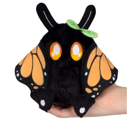 A black plush creature toy with orange and black butterfly wings, large orange eyes, and a small green leaf on its head, being held in a person's hand against a white background. Squishable Monarch Mothman - Alter Egos Series 21