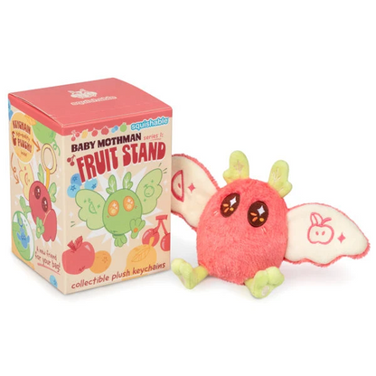 A pink plush baby moth keychain with green legs and cream wings stands next to its illustrated retail box on a white background. The box reads 'squishible BABY MOTHMAN FRUIT STAND' and shows colorful fruit-themed artwork.   Squishable Mothman Fruit Stand Blind Box -Plush Keyring Series 1