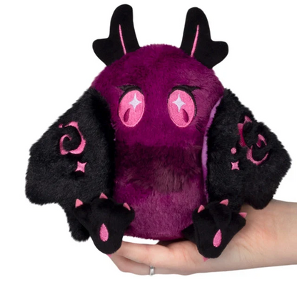 A purple and black plush toy shaped like a cute bat or creature, held in a person's hand against a plain white background. The plush has fuzzy purple body, black wings and ears, pink eye patches with star details, and pink claw accents. Squishable Poison Mothman - Alter Egos Series 21