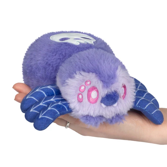 A purple plush monster toy with blue webbed gloves and pink paw-print details, being held in a person's hand against a white background. Squishable Skull Spider - Alter Ego Series 20