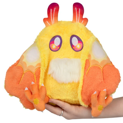 A fluffy yellow and orange plush toy with large purple eyes and small horn-like protrusions on top, being held in a person's hand against a plain white background. Squishable Solar Mothman