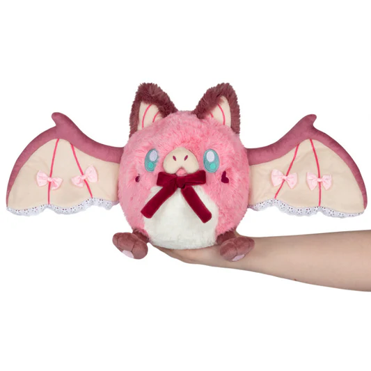 Plush toy resembling a pink bat with a red bow held by a hand on a white background. Squishable Sweetheart Bat