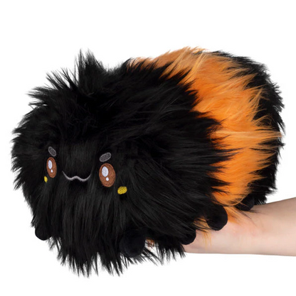 Plush toy with black and orange fur held by a hand on a white background. Squishable Woolly Caterpillar plush soft toy with fluffy black and orange fur