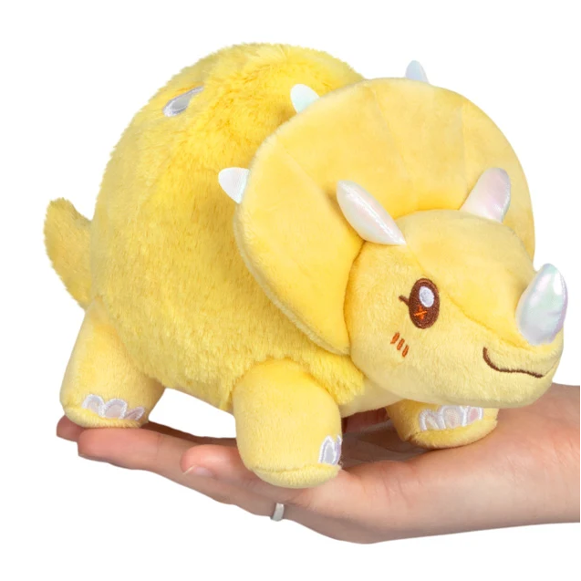A yellow plush toy triceratops dinosaur being held on a person's hand against a plain white background. Squishable Yellow Triceratops - Snackers S1E6 Limited Edition