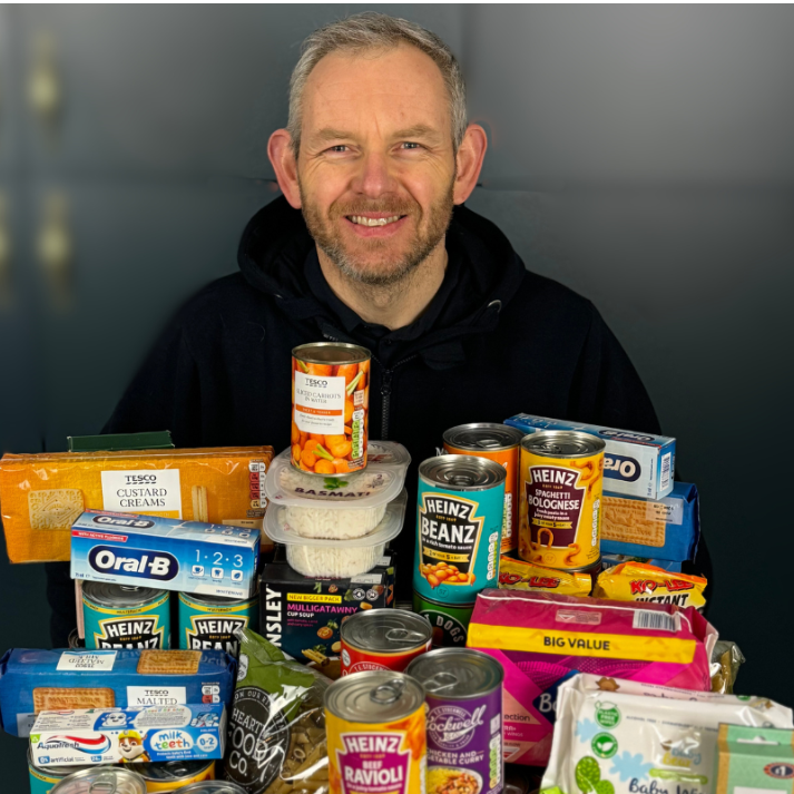 Man with a pile of food items including canned goods and boxes on a table.