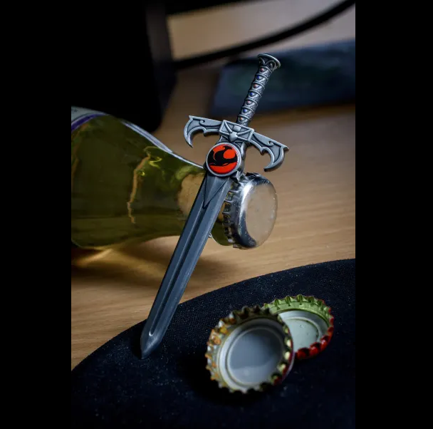 A miniature silver sword with a red and black circular emblem on the hilt, resting on a bottle cap with other bottle caps nearby on a wooden surface. ThunderCats Sword of Omens officially licensed zinc alloy bottle opener