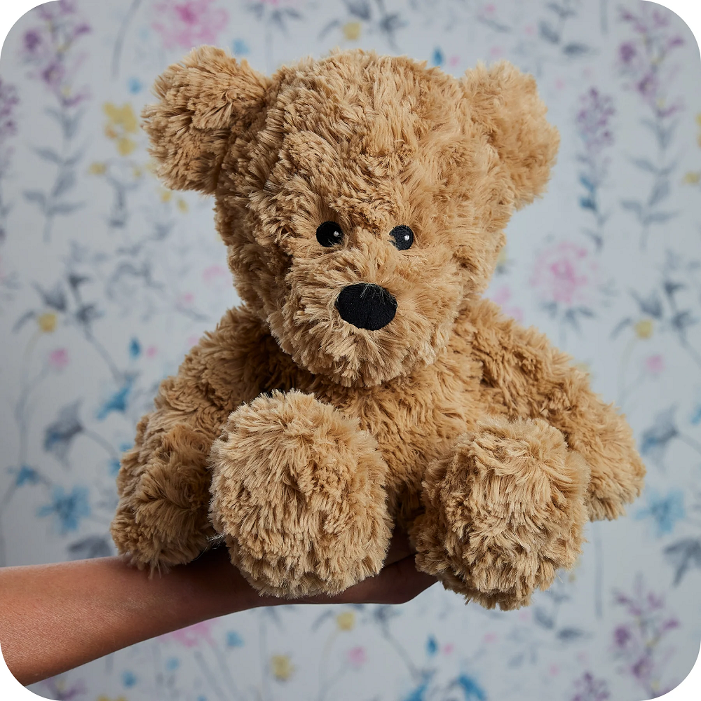 Warmies Brown Curly Bear Microwavable Lavender Plush Toy. Brown teddy bear held in a hand with a floral background