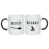 two white mugs with white inside and black handles, one mug has a picture of a broomstick and the word Witch the other has a picture of a hat and says wizard. 
