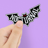 A wonderfully alternative iron on patch. This bat shaped cotton patch has red eyes and the word Nocturnal across it in white letters.. A hand holding the patch against a pink background