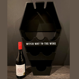 coffin shaped wine shelf is black with the words Witch Way To The Wine in white. bottle of wine on the ground next to it 